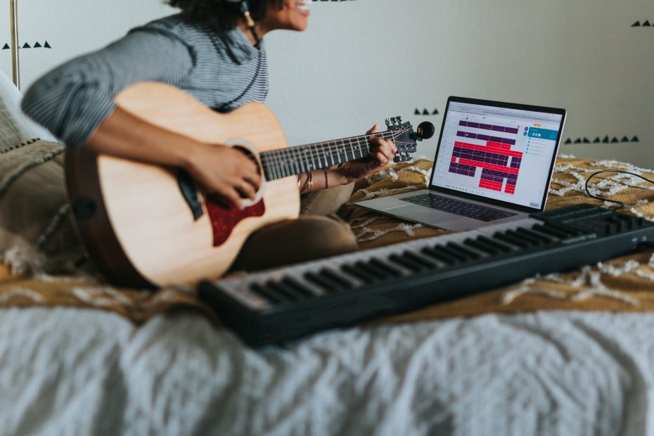 Woman sitting on a bed singing with a guitar, keyboard, and laptop.